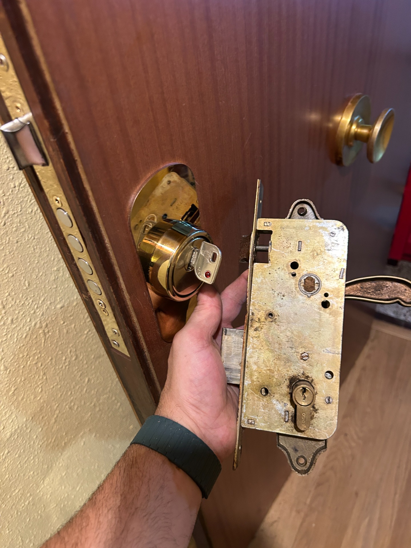 Mano sostiene una cerradura antigua de metal junto a un cilindro dorado en una puerta de madera.