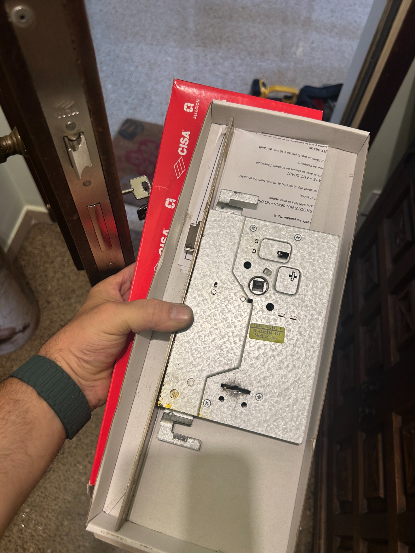 Mano sostiene una cerradura CISA, con caja roja y mecanismo metálico sobre puerta.